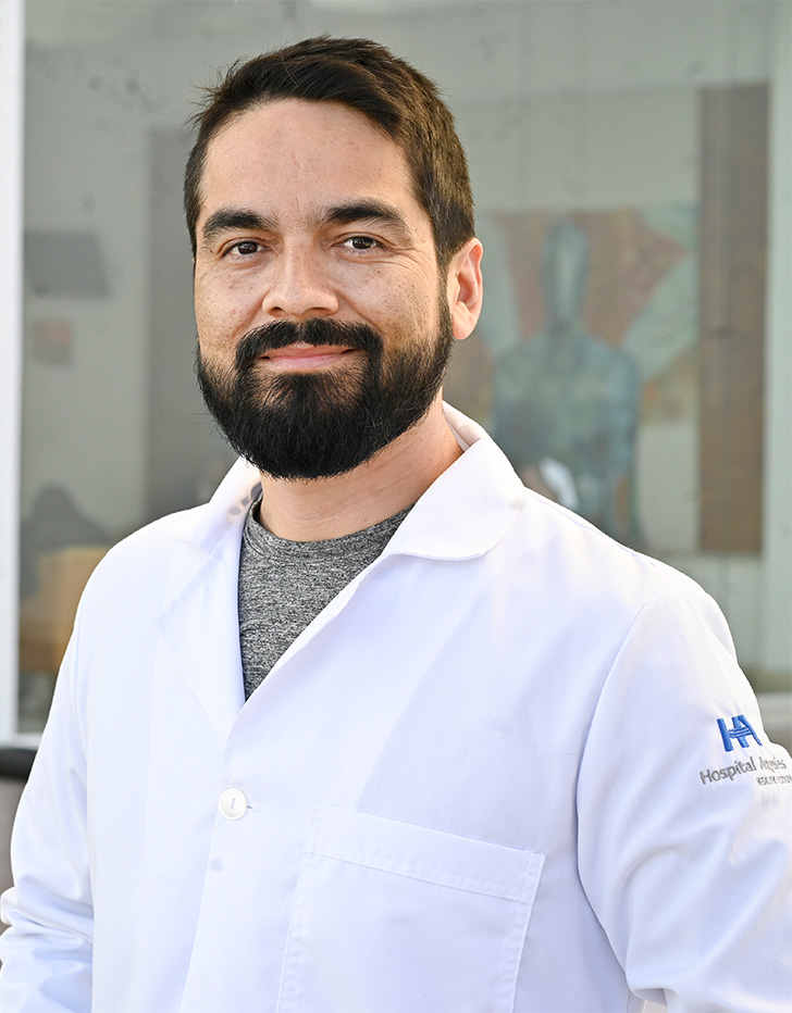 Es la foto de perfil de un urólogo en Tijuana de Hospital Angeles. Usa una bata blanca y una camisa gris. Tiene el cabello y barba café oscuro. Está sonriendo.
