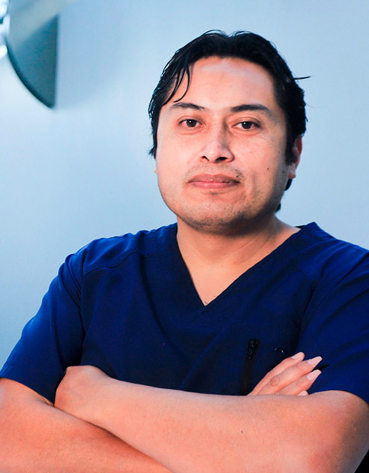 Es la foto de perfil de un cirujano general en Huixquilucan, Estado de México de Hospital Angeles Lomas. Usa un uniforme médico azul. Tiene los brazos cruzados y el cabello negro.