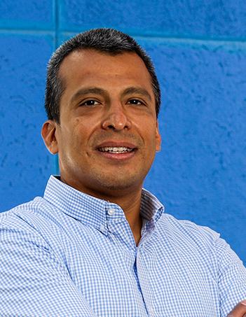 Es la foto de perfil de un endoscopista y cirujano general en León de Hospital Angeles León. Tiene una camisa azul claro, es de tez morena y cabello oscuro. El fondo es azul. 