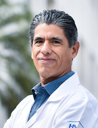 Es la foto de perfil de un ginecólogo y obstetra en Villahermosa de Hospital Angeles. Usa una bata blanca con acamisa azul. Tiene el cabello negro. Está sonriendo ligeramente.
