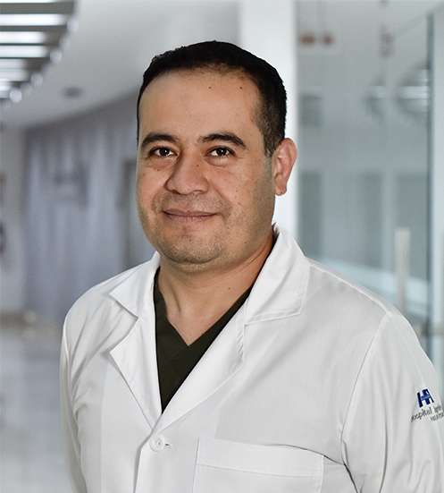 Es la foto de perfil de un angiólogo y cirujano vascular en Huixquilucan de Hospital Angeles Lomas. Usa una bata blanca con una playera negra de fondo, tiene las manos entrelazadas por detrás de la espalda y está sonriendo.