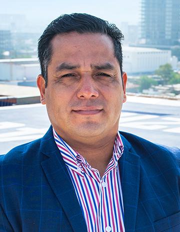 Es la foto de perfil de un urólogo en San Luis Potosí de Hospital Angeles. Usa un saco azul marino con una camisa rosa con azul de cuadros. Tiene el cabello negro. Está sonriendo ligeramente.