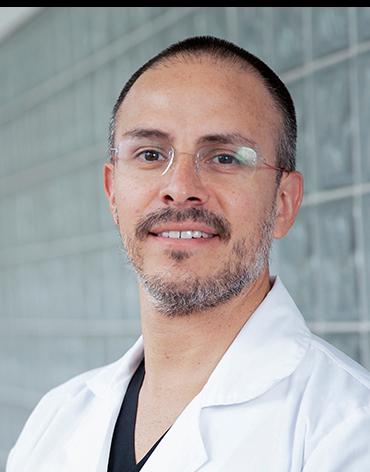 Es la foto de perfil de un oftalmólogo en Huixquilucan de Hospital Angeles Lomas. Usa una bata blanca con una playera negra debajo, el doctor usa lentes y está sonriendo.