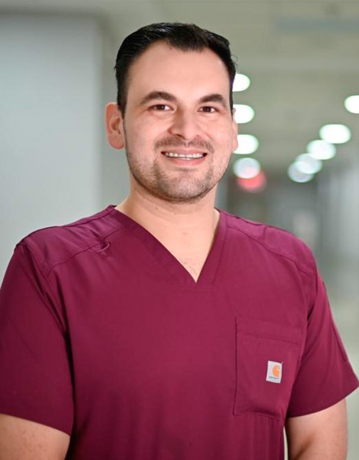 Es la foto de perfil de un ginecólogo y obstetra en Ciudad Juárez de Hospital Angeles Ciudad Juárez.  Trae puesta una bata blanca con una playera en cuello V de color azul.