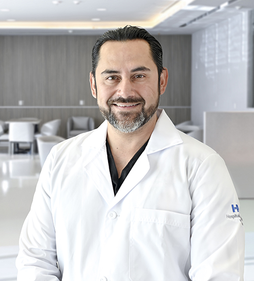 Es la foto de perfil de un cirujano general en San Pedro Garza de Hospital Angeles Valle Oriente. Usa un uniforme médico azul. Tiene el cabello y barba negro. Tiene los brazos cruzados por detrás y está sonriendo.