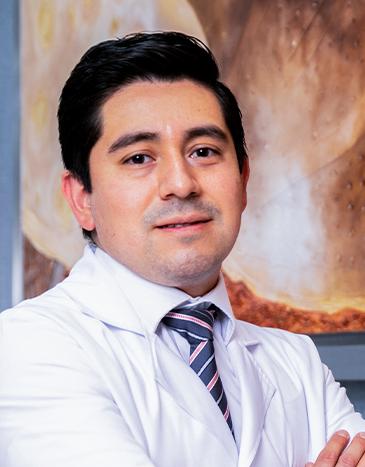 Es la foto de perfil de un urólogo en León de Hospital Angeles León. Utiliza una bata blanca con camisa blanca, corbata negra y líneas. Tiene los brazos cruzados.