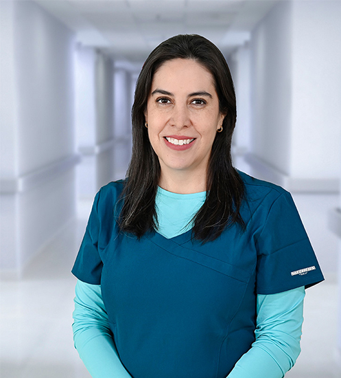 Es la foto de perfil de un odontólogo en Ciudad Juárez de Hospital Angeles Ciudad Juárez. La doctora tiene el cabello oscuro y lacio, y está sonriendo. Lleva una bata médica blanca sobre una blusa negra. En sus orejas, se pueden ver pequeños aretes de perla. El fondo muestra un área luminosa del hospital, con paredes claras y una pintura en tonos cálidos.