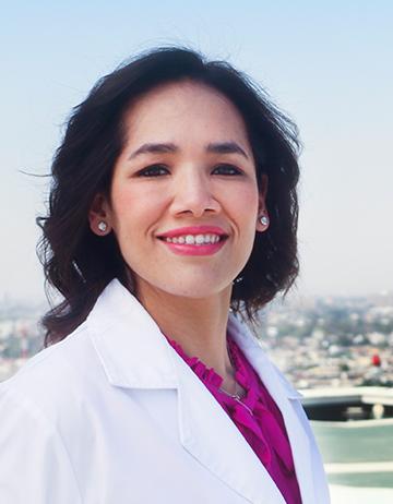 Es la foto de perfil de una endoscopista y gastroenteróloga en Torreón de Hospital Angeles Torreón. Usa una bata blanca con un uniforme rosa. Tiene el cabello corto negro y ondulado. Usa labial rojo y está sonriendo.