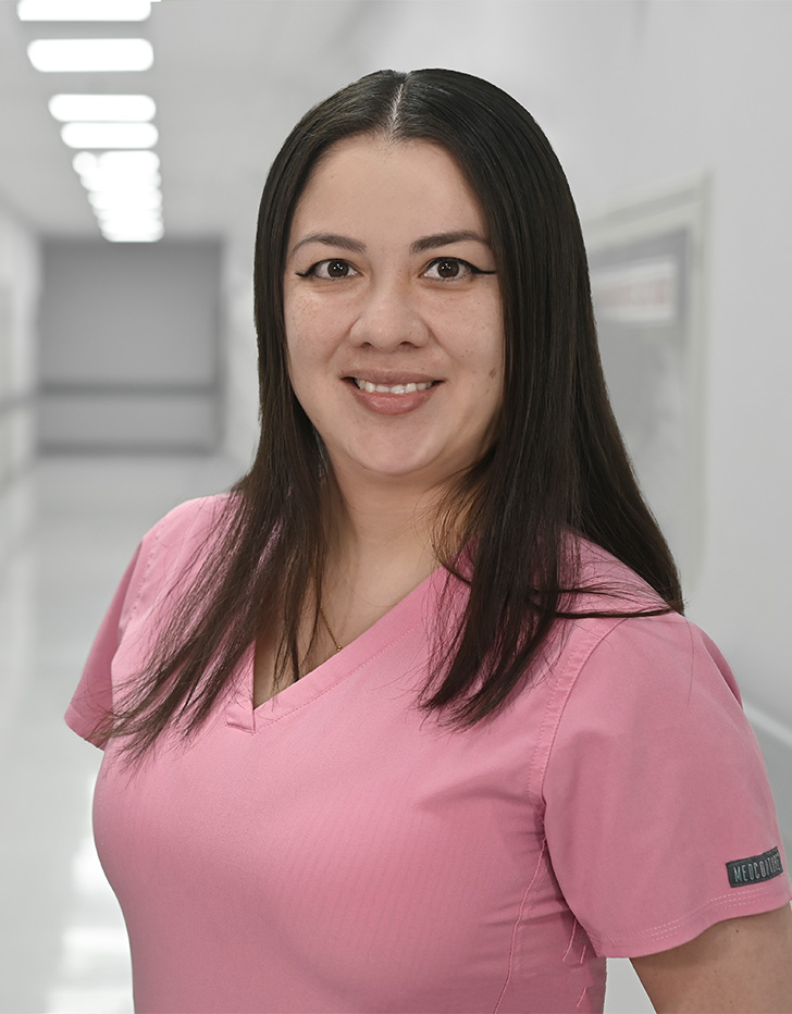 Es la foto de perfil de un pediatra en Ciudad de México de Hospital Angeles Universidad. Usa un uniforme médico color rosa. Tiene el cabello lacio y color café oscuro. Está sonriendo y tiene los brazos cruzados por detrás.