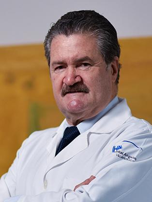 Es la foto de perfil de un cirujano general en Villahermosa de Hospital Angeles. Usa una bata blanca, camisa blanca y una corbata negra. Tiene el cabello y bigote negro. Tiene los brazos cruzados.