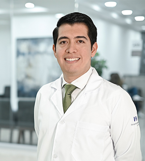 Es la foto de perfil de un cirujano general en Ciudad de México de Hospital Angeles Acoxpa. Usa una camisa y bata blanca con una corbata color verde. Está sonriendo y tiene la ceja poblada.