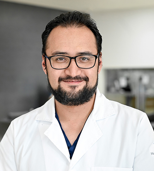 Es la foto de perfil de un cardiólogo intervencionista en San Luis Potosí. Usa una bata blanca. Tiene el cabello y barba negro. Usa unos lentes, Está sonriendo ligeramente.