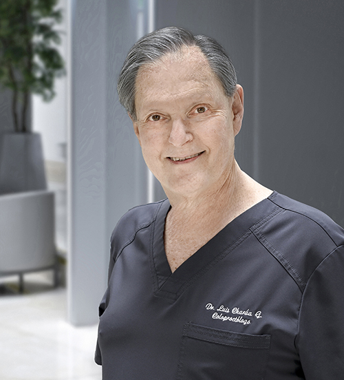 Es la foto de perfil de un coloproctólogo en Huixquilucan de Hospital Angeles Lomas. Utiliza una bata blanca con una camisa, tiene los brazos cruzados, cabello corto y canoso.