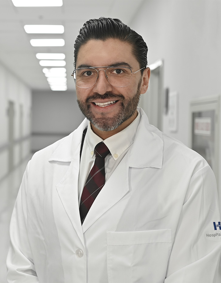 Es la foto de perfil de un urólogo en Ciudad de México de Hospital Angeles Santa Mónica y Hospital Angeles Universidad. Usa una bata blanca con una camisa blanca y una corbata vino. Tiene el cabello y barba negro.