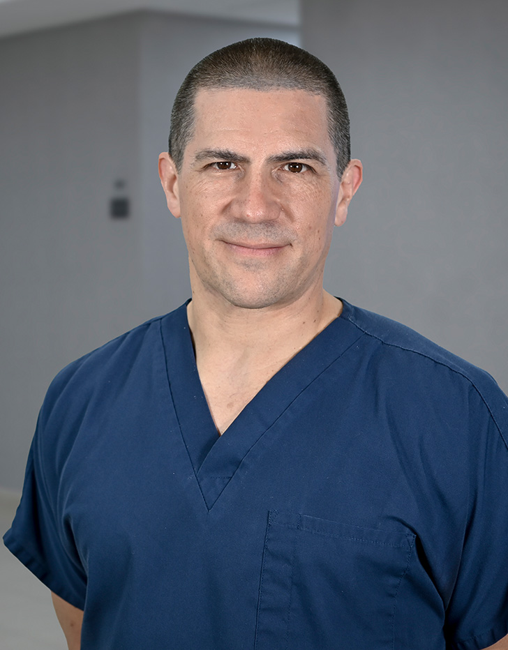 Es la foto de perfil de un cirujano general en Ciudad de México de Hospital Angeles Universidad. Usa un uniforme médico azul. Tiene el cabello muy corto y de color negro. Está sonriendo. Tiene los brazos cruzados.