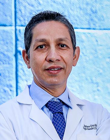Es la foto de perfil de un angiólogo y cirujano vascular en León de Hospital Angeles León. Utiliza una bata blanca, camisa azul y corbata azul. El fondo es una pared de color azul claro.