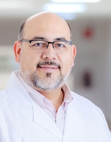 Es la foto de perfil de un cirujano pediátrico en San Luis Potosí de Hospital Angeles. Usa una bata y camisa blanca. Tiene el cabello y barba blanca y gris. Usa lentes