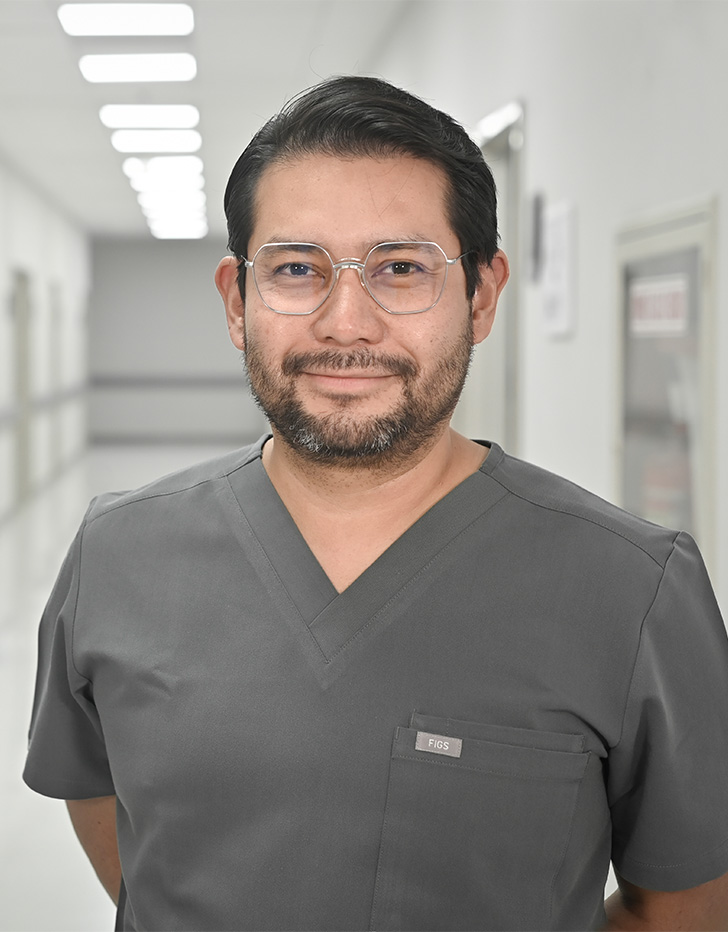 Es la foto de perfil de un cirujano general en Ciudad de México de Hospital Angeles Universidad. Usa un uniforme médico gris. Tiene el cabello y barba negra. Usa lentes, está sonriendo y tiene los brazos cruzados por detrás.