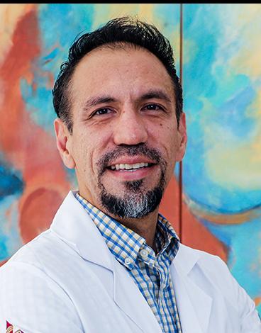 Es la foto de perfil de un nefrólogo e internista en Tampico de Hospital Angeles. Usa una bata blanca con camisa de cuadros azul y blanco. Tiene el cabello negro y barba negro con gris. Está sonriendo.