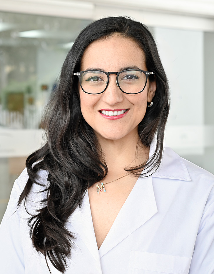 Es la foto de perfil de una psiquiatra en Tijuana de Hospital Angeles. Usa una bata blanca con un sueter negro. Tiene el cabello largo, ondulado y café oscuro. Está sonriendo.