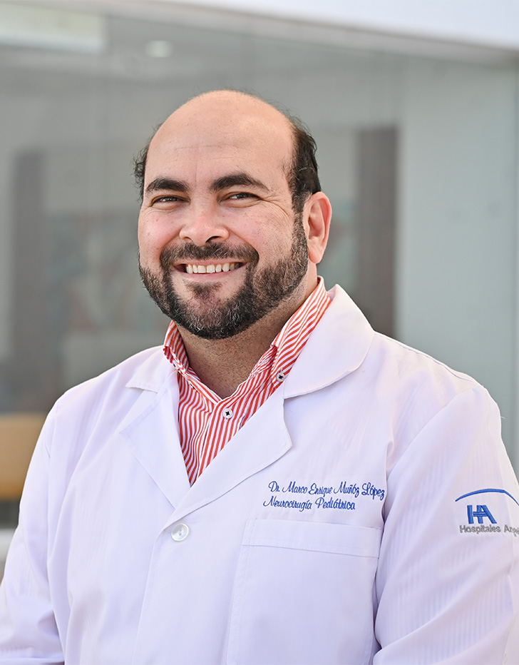 Es la foto de perfil de un cirujano neurológico y cirujano neurológico pediátrico en Tijuana de Hospital Angeles. Usa una bata blanca con una camisa color salmón. Tiene la barba y parte del cabello café oscuro. Está sonriendo.