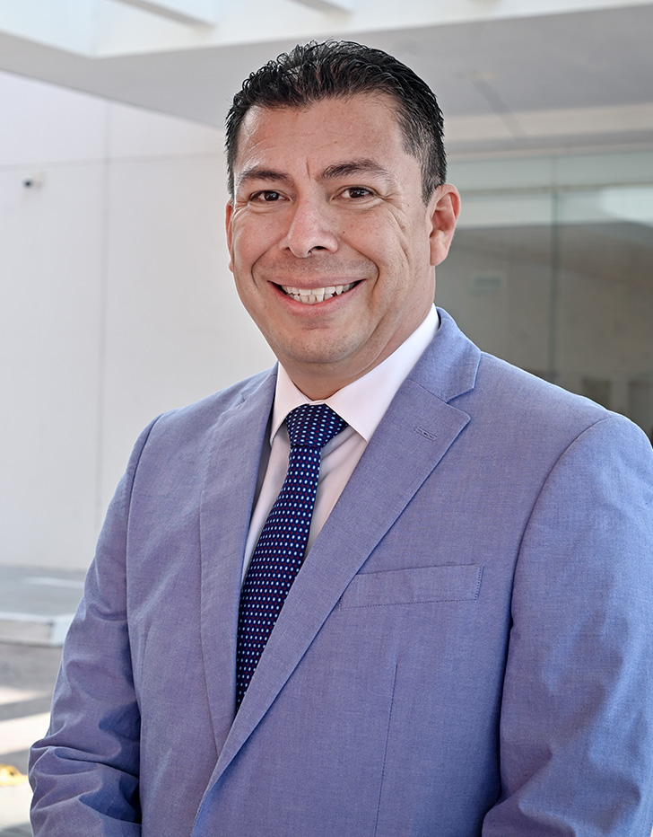 Es la foto de perfil de un otorrinolaringólogo en León de Hospital Angeles León. Utiliza una bata blanca con una camisa roja de cuadros azul marino, el doctor usa brackets y está sonriendo.