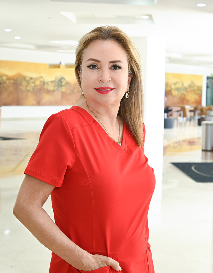 Es la foto de perfil de un especialista en audiología y foniatría en Ciudad Juárez de Hospital Angeles Ciudad Juárez. Trae un uniforme rojo y tiene el cabello largo y rubio. Utiliza una bata blanca, una blusa azul y tiene cabello negro.