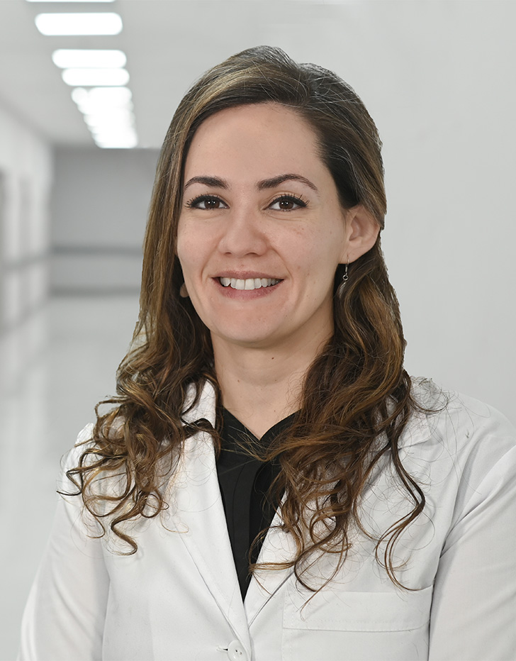 Es la foto de perfil de una uróloga en Ciudad de México de Hospital Angeles Universidad. Usa una bata blanca con un uniforme médico negro. Tiene el cabello largo,ondulado y castaño. Está sonriendo.