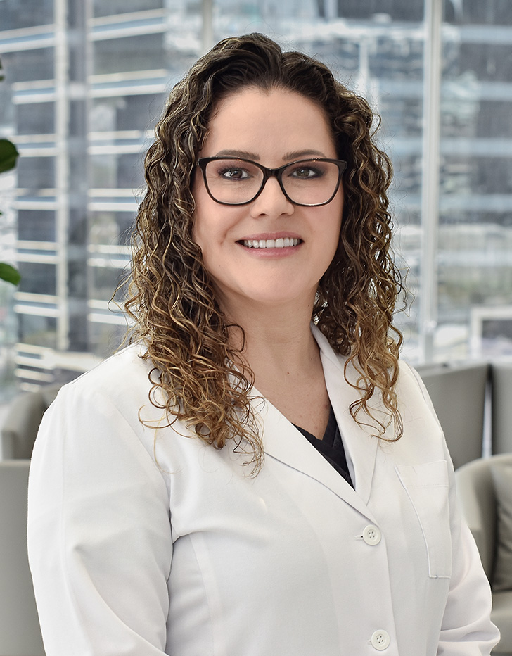 Es la foto de perfil de una médica internista en San Pedro Garza de Hospital Angeles Valle Oriente. Usa una bata blanca con un uniforme médico azul. Tiene el cabello castaño. Usa lentes y está sonriendo.