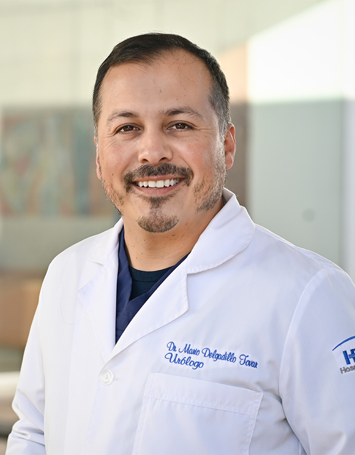 Es la foto de perfil de un urólogo en Tijuana de Hospital Angeles. Usa una bata blanca con camisa negra. Tiene el cabello y la barba gris con negro. Está sonriendo y tiene los brazos cruzados por detrás.