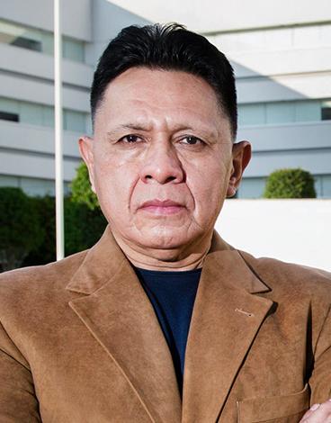 Es la foto de perfil de un artroscopista, ortopedista y traumatólogo pediátrico en Ciudad de México de Hospital Angeles. El doctor viste un saco color café claro, una playera negra y de fondo se encuentra el hospital. 