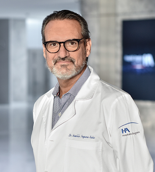 Es la foto de perfil de un ortopedista y traumatólogo en Querétaro de Hospital Angeles Centro Sur. El doctor tiene una bata blanca y una camisa gris tiene barba canosa y lentes con marco negro.