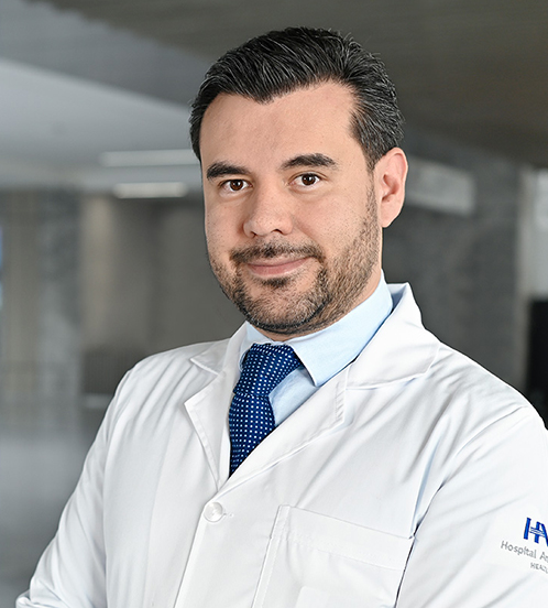 Es la foto de perfil de un urólogo en Querétaro de Hospital Angeles Centro-sur. Usa una bata blanca con una camisa azul claro y una corbata color azul oscuro y puntos blancos.