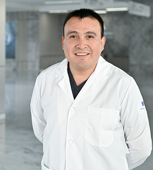 Es la foto de perfil de un ortopedista y traumatólogo pediatra en Querétaro de Hospital Angeles Centro-sur. Usa una bata blanca con uniforme médico azul oscuro. Tiene el cabello negro, está sonriendo y sus manos están por detrás.