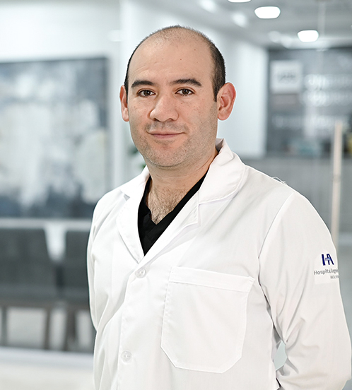 Es la foto de perfil de un cirujano oncólogo en Ciudad de México de Hospital Angeles Acoxpa. Usa una bata  color blanca y un uniforme médico color azul oscuro. Tiene muy poco cabello y los brazos cruzados atras.