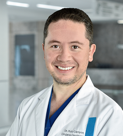 Es la foto de perfil de un cirujano oncológico en Querétaro de Hospital Angeles Centro Sur. Viste una bata blanca y una camisa azul está sonriendo.