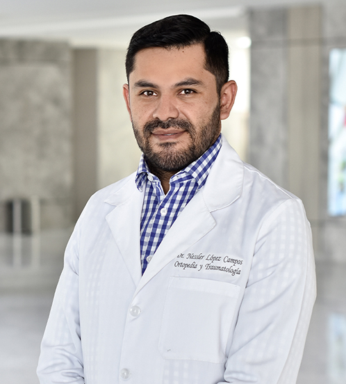 Es la foto de perfil de un ortopedista y traumatólogo en Querétaro de Hospital Angeles Centro Sur. El doctor tiene una bata blanca con una camisa azul con cuadros blancos el doctor tiene un uniforme negro y está sonriendo.