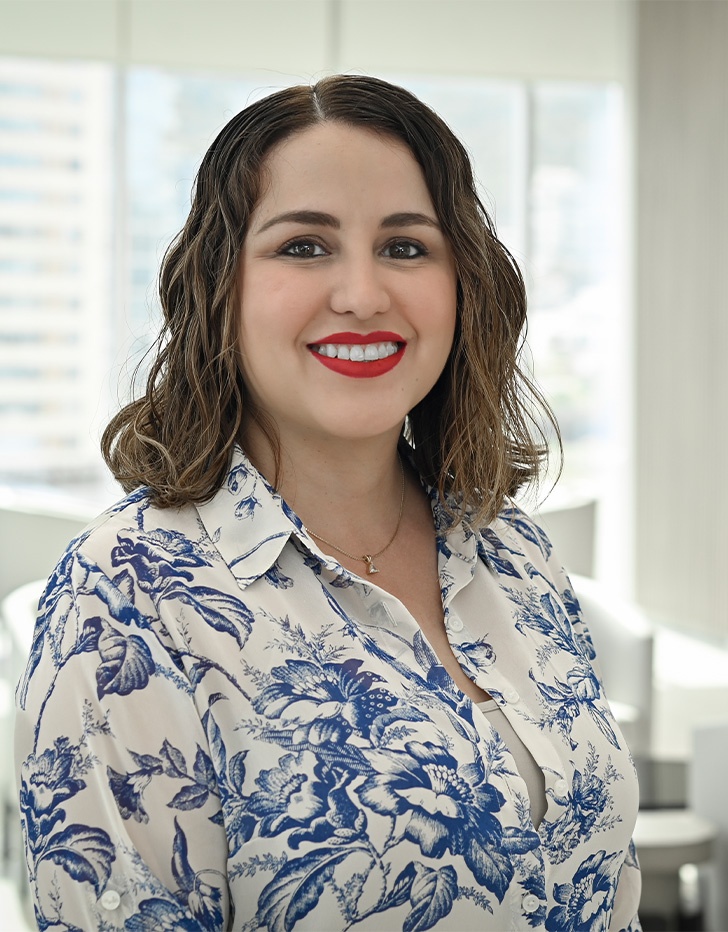 Es la foto de perfil de una ginecóloga y obstetra en San Pedro Garza de Hospital Angeles Valle Oriente. Usa una bata blanca con flores azules. Tien el cabello corto, ondulado y castaño. Está sonriendo y usa un labial rojo.