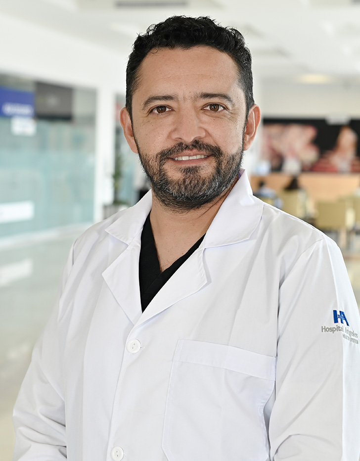 Es la foto de perfil de un cirujano oncológico y cirujano general en Xalapa de Hospital Angeles Xalapa. Usa una bata blanca con un uniforme médico azul. Tiene el cabello y barba negra. Tiene los brazos cruzados.