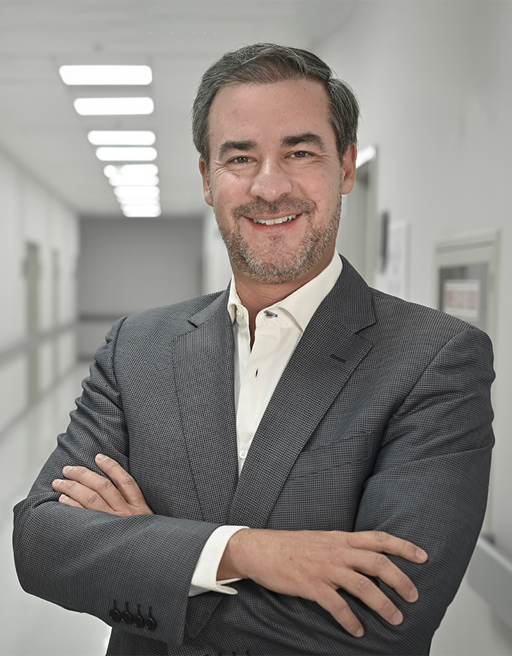 Es la foto de perfil de un urólogo en Ciudad de México de Hospital Angeles Universidad. Usa un saco gris y camisa blanca. Tien el cabello y barba negro con gris. Está sonriendo y tiene los brazos cruzados.