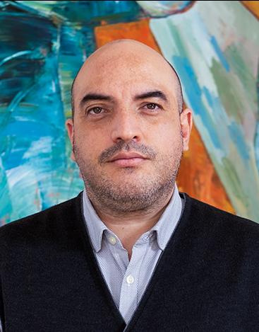 Es la foto de perfil de un reumatólogo en Tijuana de Hospital Angeles Tijuana. Usa un sueter negro con una camisa azul. Es calvo y está serio.