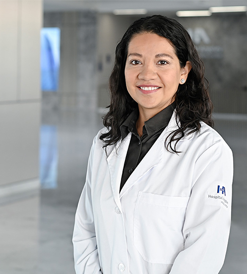 Es la foto de perfil de una cirujana general en Querétaro de Hospital Angeles Centro-sur. Usa una bata blanca con una blusa negra. Tiene el cabello corto y ondulado. Sus brazos están cruzados en la parte de enfrente.