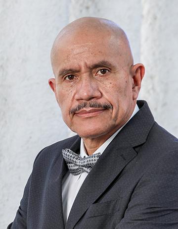 Es la foto de perfil de un hematólogo en León de Hospital Angeles León. Usa un saco negro con camisa blanca y un moño negor con blanco. El doctor es calvo y tiene bigote.