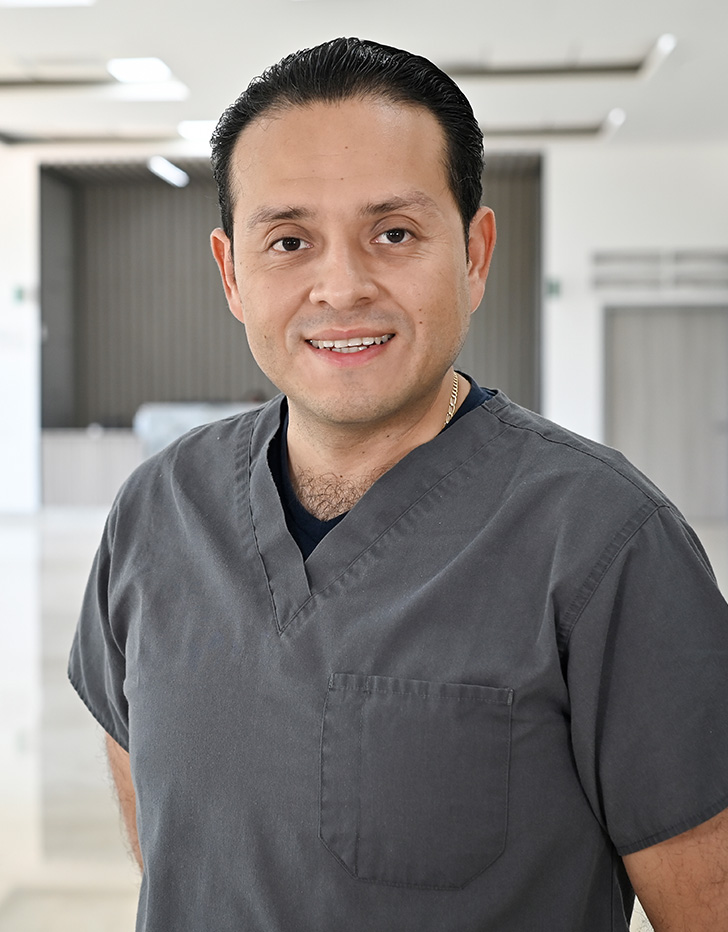 Es la foto de perfil de un pediatra en Tampico de Hospital Angeles Tampico. Usa una bata blanca con una camisa blanca. Usa un moño negro en el cuello. Tiene el cabello negro y está sonriendo.