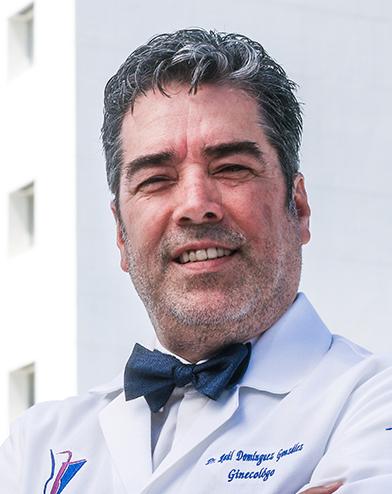 Es la foto de perfil de un ginecólogo y obstetra en Torreón de Hospital Angeles Torreón. Usa una bata blanca con camisa blanca y moño azul. Tiene el cabello gris y la barba blanca. Está sonriendo.