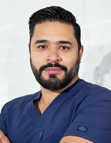 Es la foto de perfil de un urólogo en Ciudad de México de Hospital Angeles Universidad. Usa un uniforme médico azull marino. Tiene el cabello y la barba negra. Tiene los brazos cruzados.