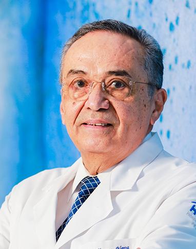 Es la foto de perfil de un cardiólogo intervencionista en Ciudad de México de Hospital Angeles Pedregal. Usa una bata blanca con una camisa blanca y una corbata azul. Tiene el cabello blancon con gris. Está sonriendo ligeramente.