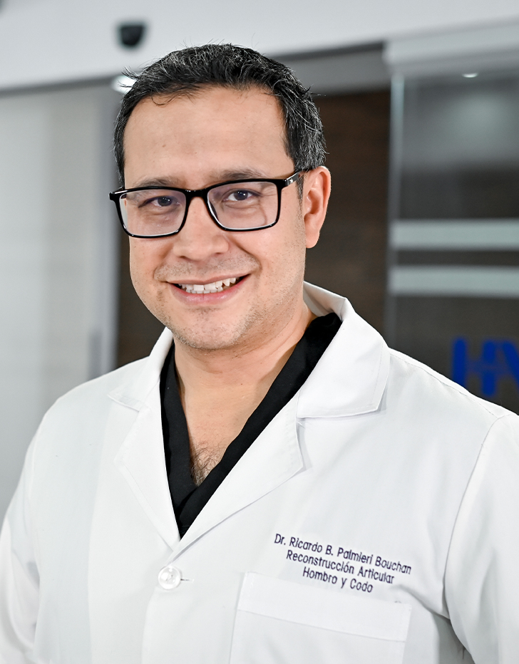Es la foto de perfil de un ortopedista y traumatólogo en Ciudad de México de Hospital Angeles Santa Mónica. Usa una bata blanca con un uniforme médico negro. Tiene el cabello negro. Usa lentes y está sonriendo.