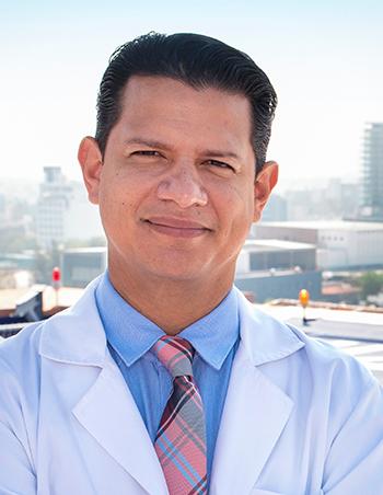 Es la foto de perfil de un especialista en angiología y cirugía vascular y endovascular en San Luis Potosí de Hospital Angeles San Luis Potosí. Usa una bata blanca con camisa azul y corbata de colores. Tiene el cabello negro.