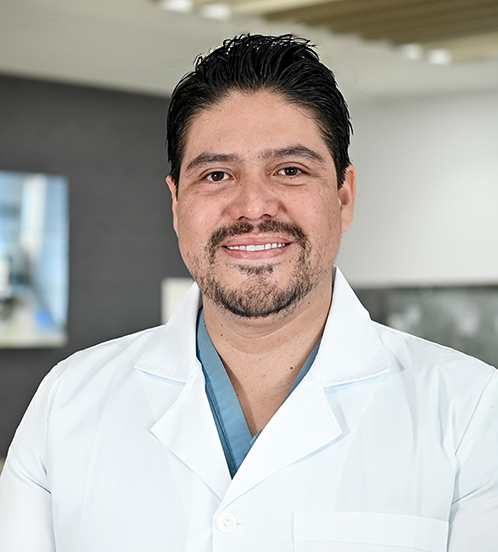 Es la foto de perfil de un ginecólogo y obstetra en San Luis Potosí de Hospital Angeles San Luis Potosí. Usa una bata blanca con un uniforme médico azul. Tiene el cabello y barba negra. Está sonriendo.